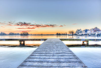 Pier amidst frozen lake against sky during sunset