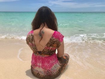 Rear view of tattooed woman sitting at beach against sky on sunny day