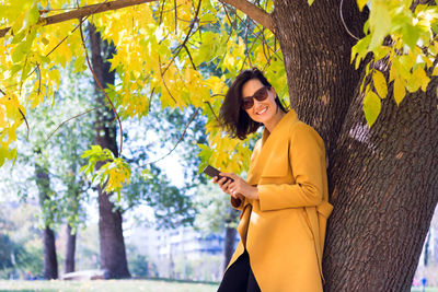 Happy woman in yellow coat using smart phone in park in autumn.