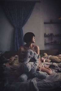 Naked woman sitting with stuffed toy on bed at home