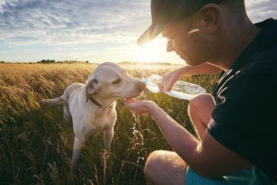 Man feeding water to dog on field against sky