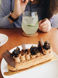 Cropped image of woman sitting by chocolate eclair and drink served on table