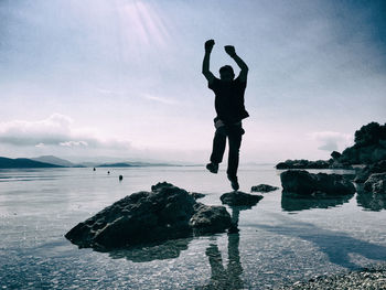 Full length of man with arms raised jumping at beach against sky