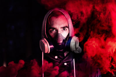 Portrait of young man wearing gas mask amidst red smoke at night