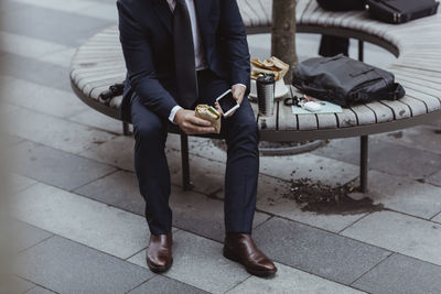 Low section of entrepreneur with smart phone and sandwich sitting in city