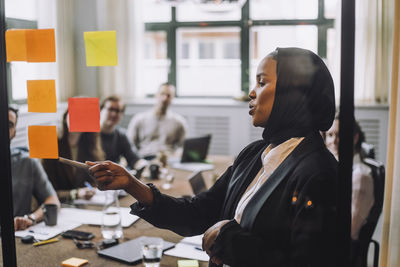 Young businessman wearing headscarf explaining adhesive notes to colleagues during meeting in creative office