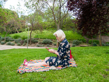 Full length of mature woman using mobile phone while sitting on picnic blanket in park