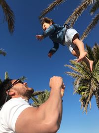 Low angle view of man playing while throwing boy against trees and clear blue sky