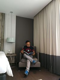 Full length of man reading book while sitting on chair in hotel room