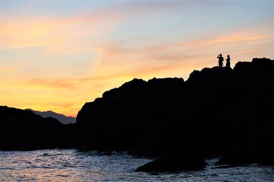 Silhouette people on rocks by sea against sky during sunset