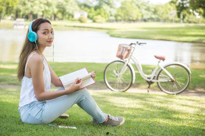 Portrait of young woman holding book while listening music on grass by pond
