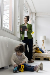 Young businesswoman standing at window working, whilde daughgter is playing on floor