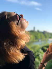 Side view of young woman eating ice cream cone while standing against blue sky