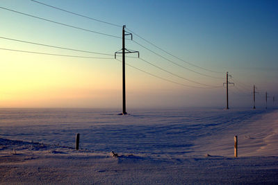 Electricity pylon on snowy field against sky during sunset