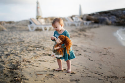Cute european baby toddler in a green dress plays with a doll on the beach by the sea at sunset