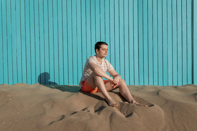 Young man with eyes closed sitting in sand beach in turquoise wall