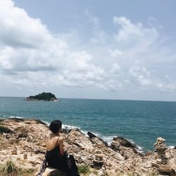 Rear view of woman sitting on rock by sea against sky