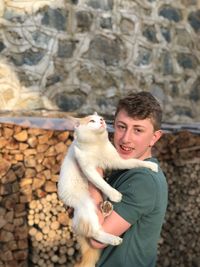 Portrait of teenage boy holding cat while standing against wall