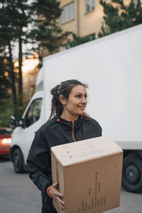 Smiling worker carrying box while standing against delivery van on street in city