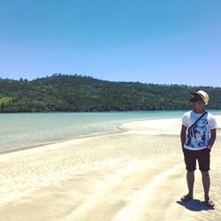 Smiling man standing on sea shore by mountain against clear blue sky