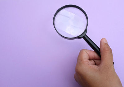 Close-up of hand holding magnifying glass against purple background