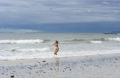 Mid adult woman in bikini standing on shore against sky