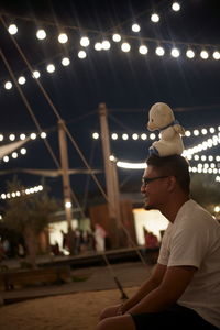 Man with teddy bear on head sitting against sky at night