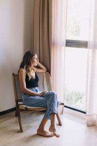 Side view of woman sitting on chair looking through the window at home