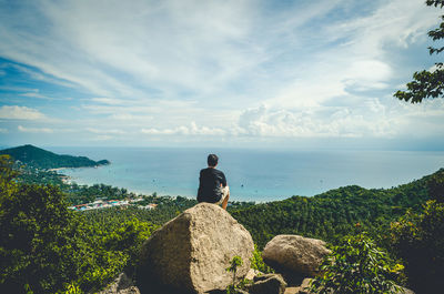 Rear view of man sitting on rock by sea against sky