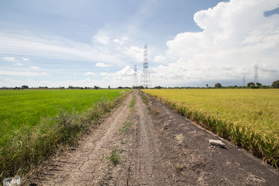 Dirt road passing through field against cloudy sky