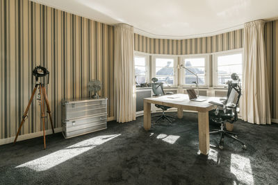 Wooden table with laptop and comfortable armchairs placed near panoramic window in spacious room with striped wallpaper in contemporary apartment