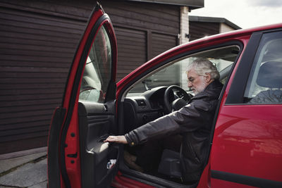 Senior man sitting in red car and closing door against garage