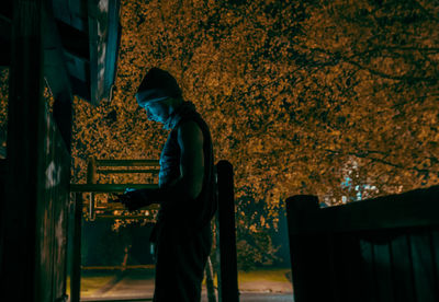 Side view of man using mobile phone by tree at night