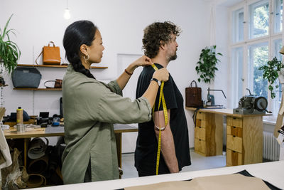 Side view of female fashion designer taking measurement of customer standing at workshop