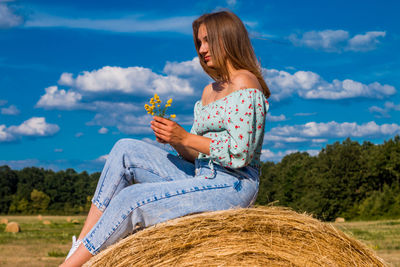 Young woman sitting on hay bale with flowers in her hands 