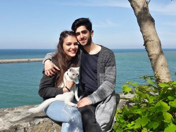 Portrait of smiling young couple with dog sitting on retaining wall against sky