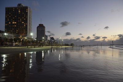 Illuminated buildings by river against sky at sunset