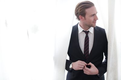 Thoughtful businessman adjusting necktie while standing amidst white curtains