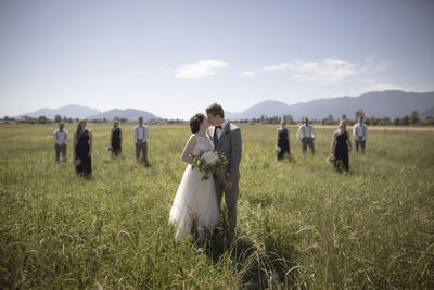 Newlywed couple kissing while standing at grassy field with groomsmen and bridesmaids in background during sunny day