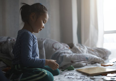 Side view of girl playing while sitting on bed at home