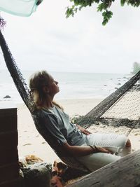 Mid adult woman looking away while relaxing on hammock at beach against sky