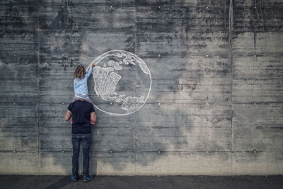 Austria, salzburg, father with daughter on his shoulders, the daughter draws with chalk the earth on a concrete wall