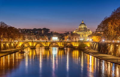 Illuminated arch bridge over river by buildings against sky