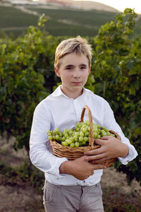 Teenage boy in a white shirt stands in a vineyard at sunset and holds a basket of green grapes