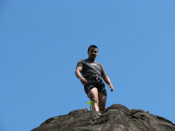 Low angle view of young man standing on rock formation against clear blue sky