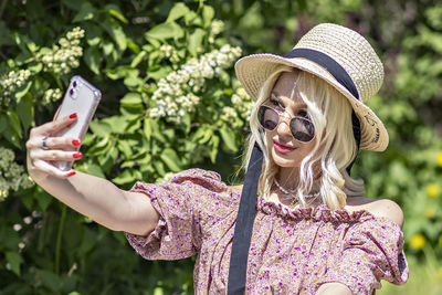 A young blonde woman in a hat and sunglasses at a picnic in the park .takes a selfie on a smartphone