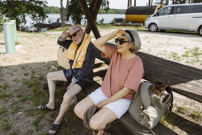 Retired senior couple sitting on bench with backpack during sunny day