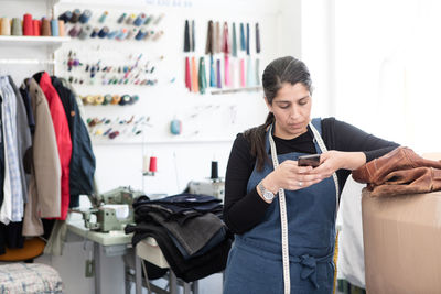 Mature female owner standing by cardboard box while using mobile phone at laundromat