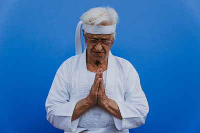 Senior woman in karate uniform with hands clasped against blue background