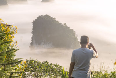 Rear view of man photographing through camera standing on mountain during foggy weather